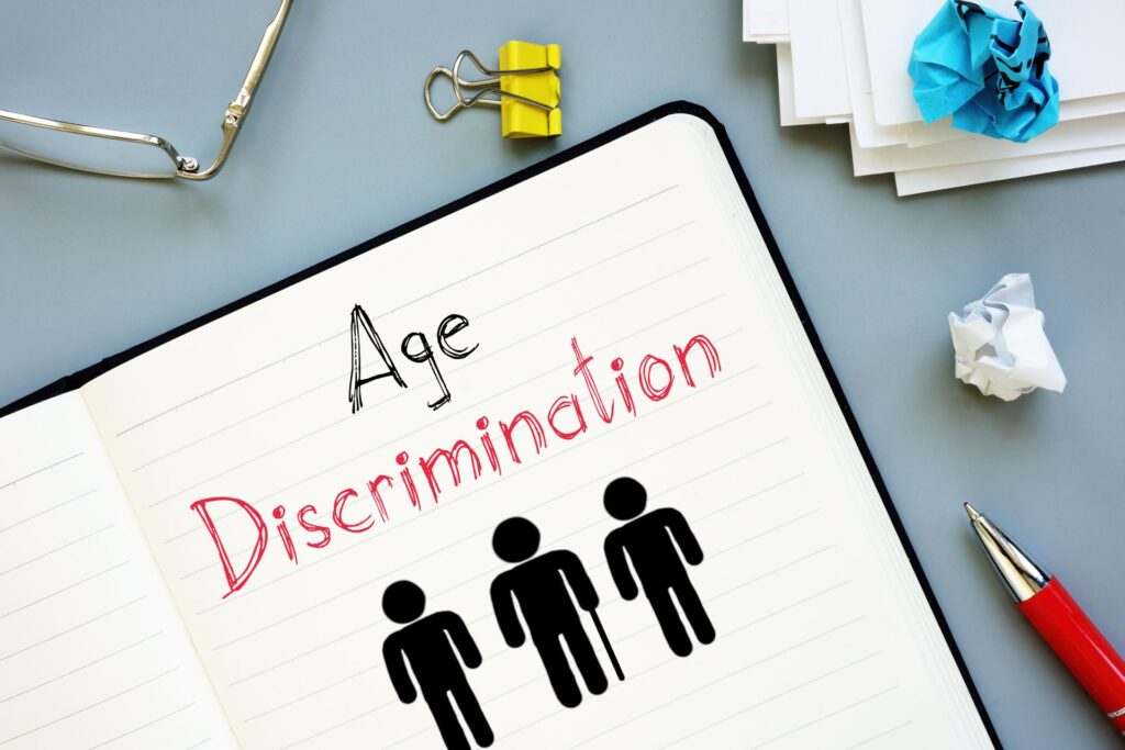 Age Discrimination is written in a notebook next to a pen, glasses and crumbled paper