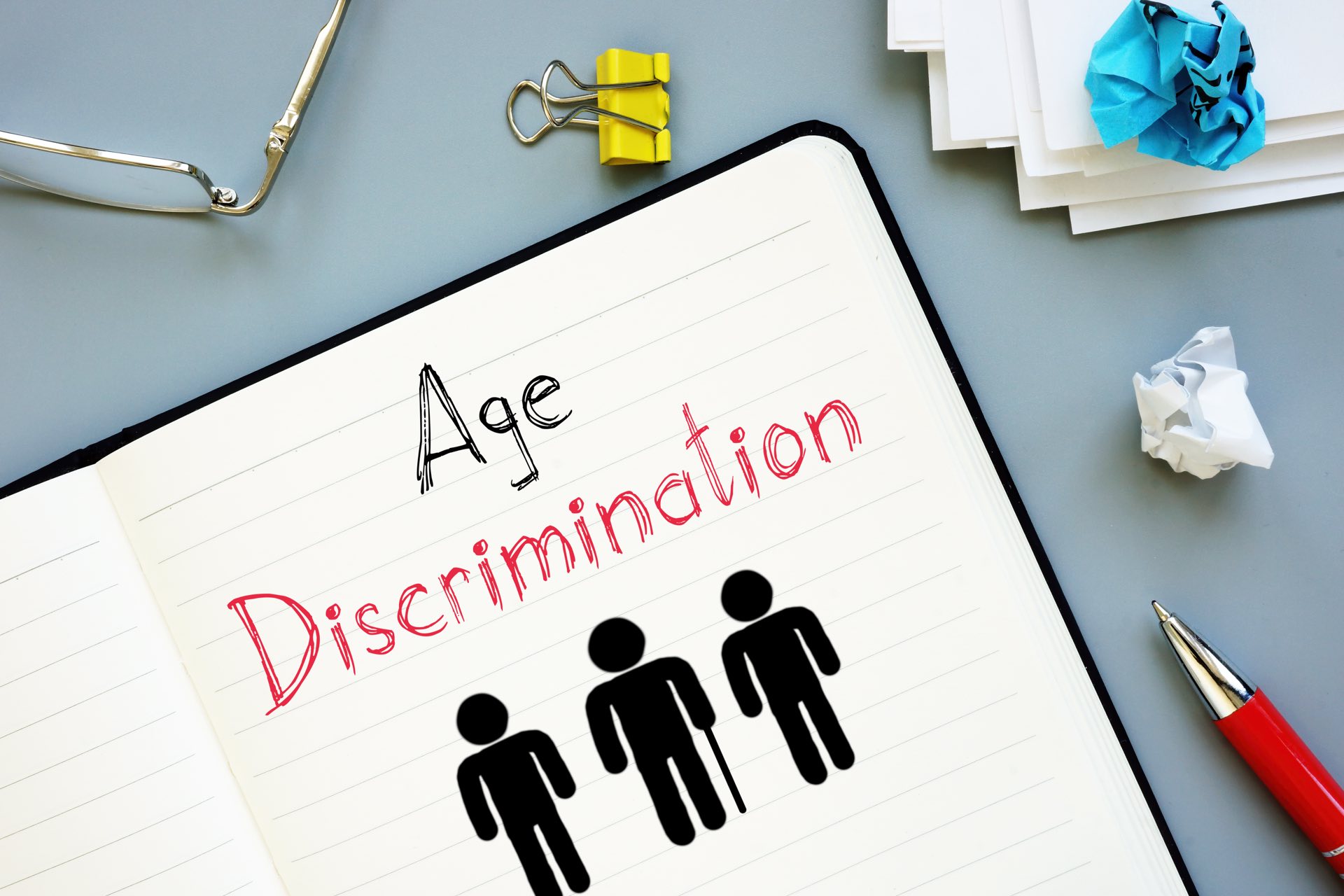 Age Discrimination is written in a notebook next to a pen, glasses and crumbled paper