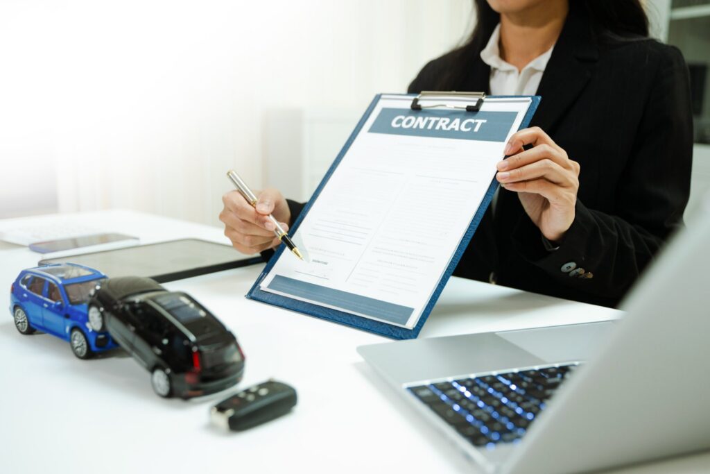 Two toy cars on a desk next to a laptop and a person holding a contract on a clipboard
