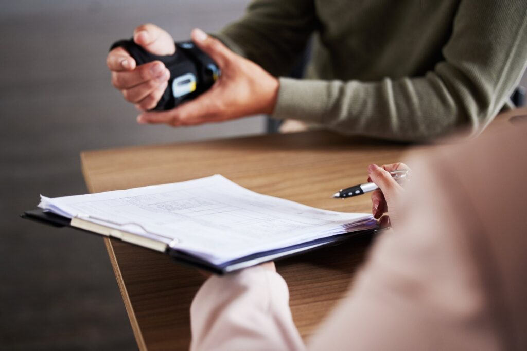 Two people sitting at a desk. One is holding a clipboard and paperwork while the other is holding his wrist