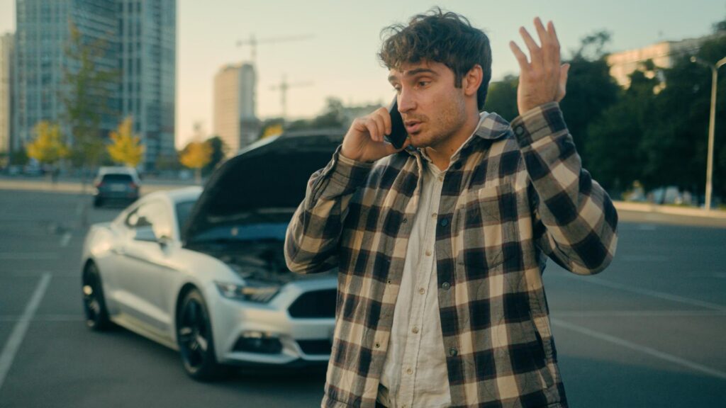 A man in a plaid jacket stands in the road talking on a cell phone in front of a car crash.