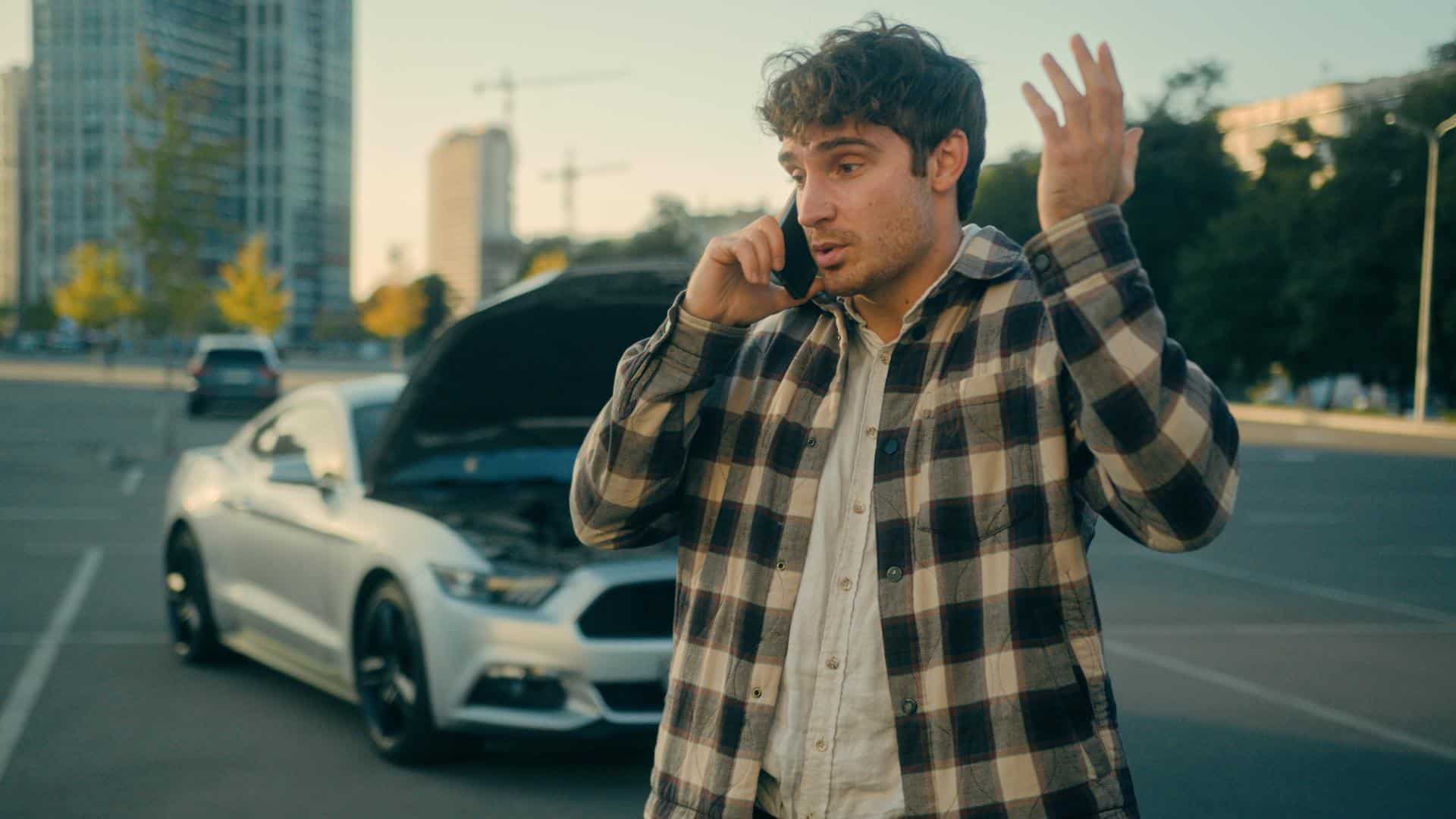 A man in a plaid jacket stands in the road talking on a cell phone in front of a car crash.