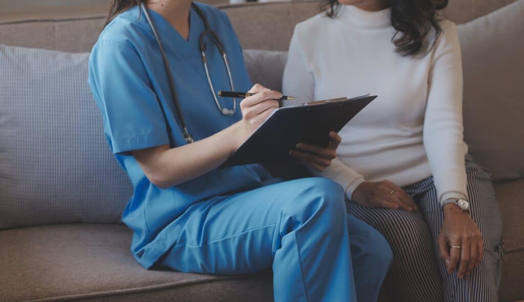 Person in blue scrubs sitting on a couch holding a clipboard going over it with another person sitting on the couch