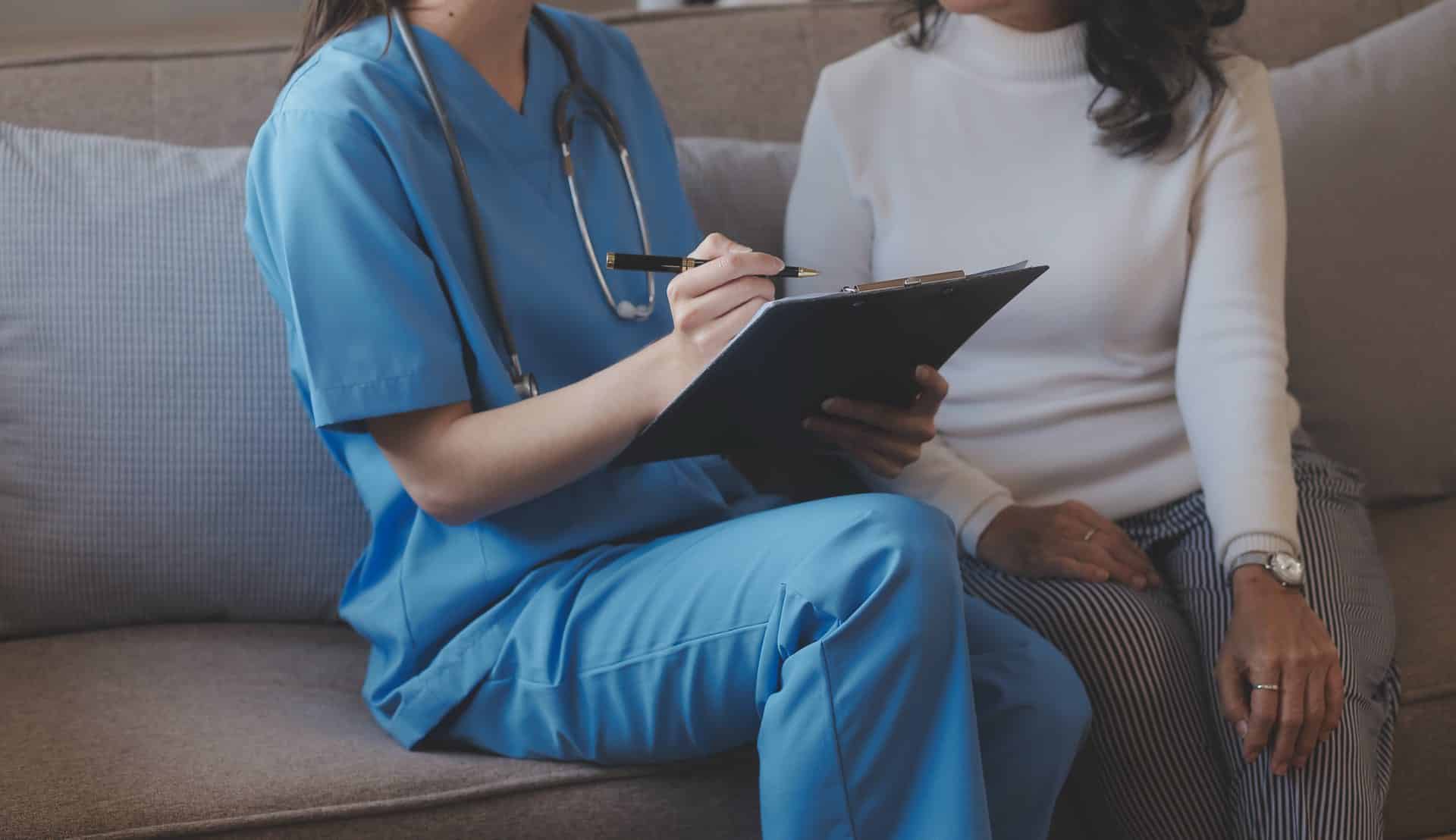 Person in blue scrubs sitting on a couch holding a clipboard going over it with another person sitting on the couch