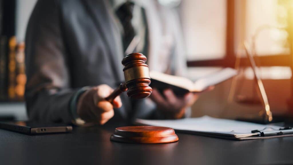 A lawyer in a business suit sits behind a desk holding a gavel and a law book.