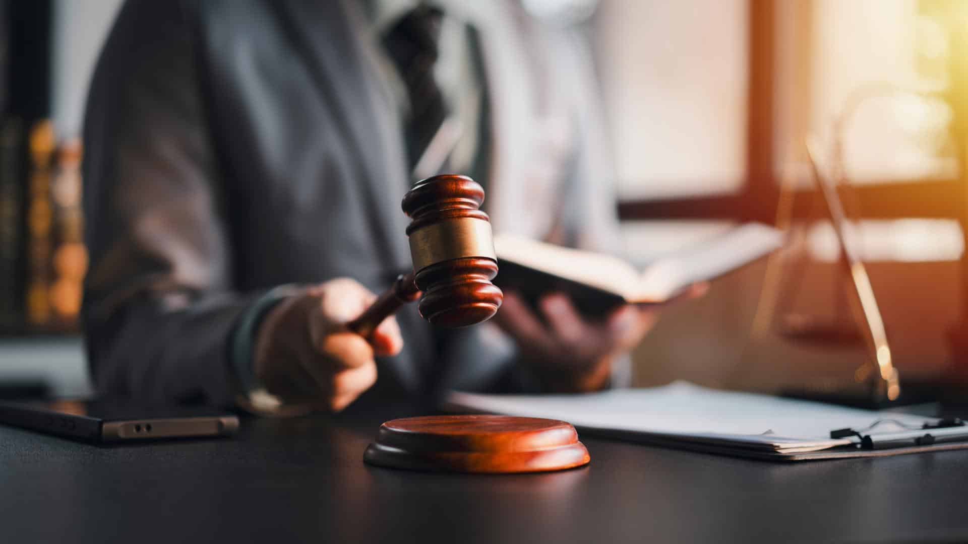 A lawyer in a business suit sits behind a desk holding a gavel and a law book.