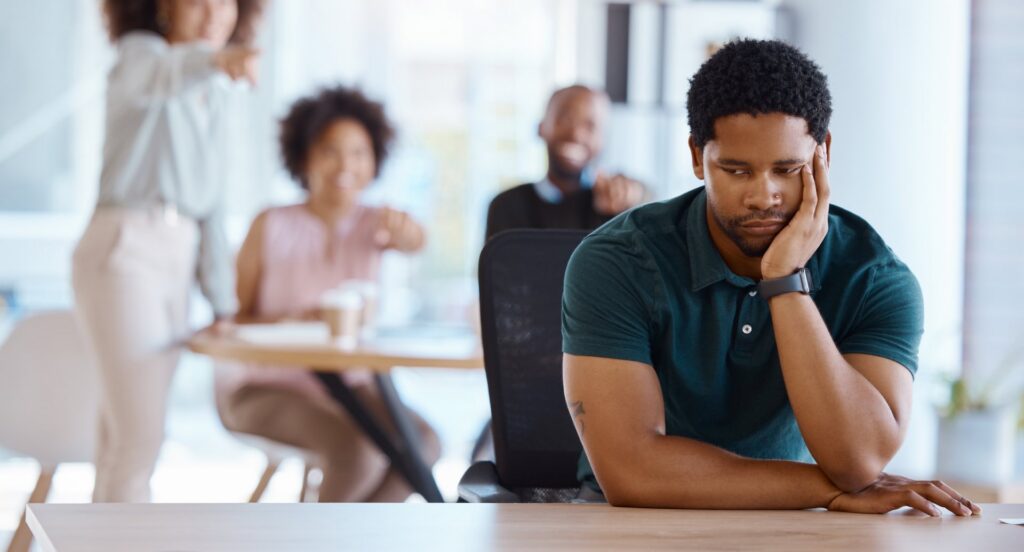 People sitting at a desk blurred in the background pointing and laughing at a person sitting in front of them at a desk