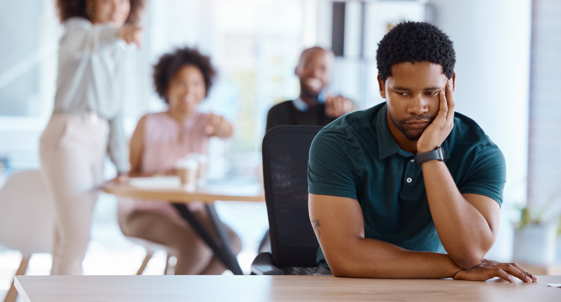 People sitting at a desk blurred in the background pointing and laughing at a person sitting in front of them at a desk