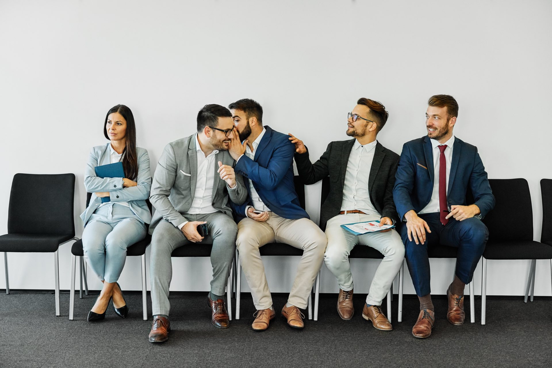 Group of young businessmen and women sitting in office chairs and one whispering to another
